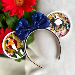 May include: White headband with a blue bow and two white ears. Each ear features a 3D design of a scene from the movie Mary Poppins Returns. One ear shows Mary Poppins riding a horse with penguins in the background. The other ear shows a man in a yellow suit with penguins in the background.