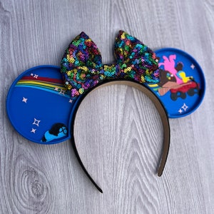 May include: Blue mouse ears headband with a rainbow and stars on one ear and a cartoon image of a character on the other ear. The headband has a large, colorful sequined bow.