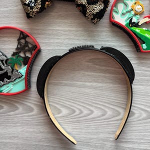 May include: Black headband with small ears, embellished with rhinestones. A gold and black sequined bow and two decorative ear-shaped accessories are also visible. The items are on a light wood-grain surface.
