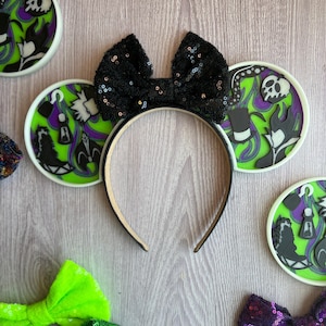 May include: A black sequined bow headband with round ear accents featuring villain-themed designs in green, purple, and black. The headband is surrounded by other decorative bows and ear accents.