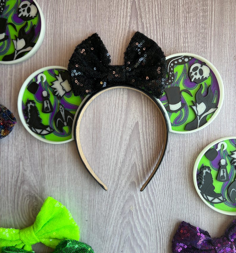 May include: A black sequined bow headband with round ear accents featuring villain-themed designs in green, purple, and black. The headband is surrounded by other decorative bows and ear accents.