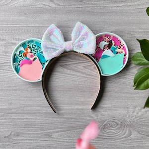 May include: A pair of light blue and pink mouse ears with a white bow. The ears feature a princess in a pink dress and a princess in a blue dress.