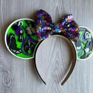 May include: A black headband with two round ear designs and a sequined bow. The ears feature a green and purple design with black silhouettes. The bow is covered in colorful sequins. The headband is a fun accessory.