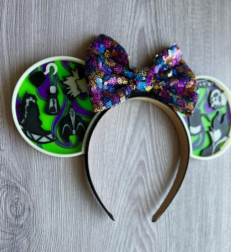 May include: A black headband with two round ear designs and a sequined bow. The ears feature a green and purple design with black silhouettes. The bow is covered in colorful sequins. The headband is a fun accessory.