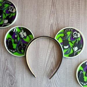 May include: Black headband with mouse ears featuring a green, purple, and black design. The ears have a white border and depict various villainous characters and symbols, including skulls, tentacles, and silhouettes.