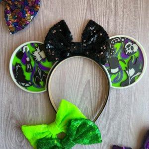 May include: A black sequined bow headband with two round ear accents featuring villain-themed designs in green, purple, and black. Additional sequined bows in green and a multi-colored sequined bow are also visible.