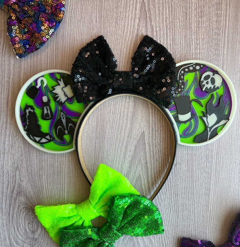 May include: A black sequined bow headband with two round ear accents featuring villain-themed designs in green, purple, and black. Additional sequined bows in green and a multi-colored sequined bow are also visible.