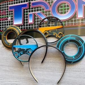 May include: A set of four black and blue Tron-themed mouse ears with a light blue, black, and yellow light cycle design, a black and blue circle design, and a black and blue design with a light cycle and a figure standing on a blue platform. The mouse ears are on a wooden surface.