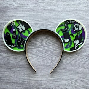 May include: A black headband with two round ear decorations. The ears feature a green and purple background with black and white character designs. The ears have a white border.
