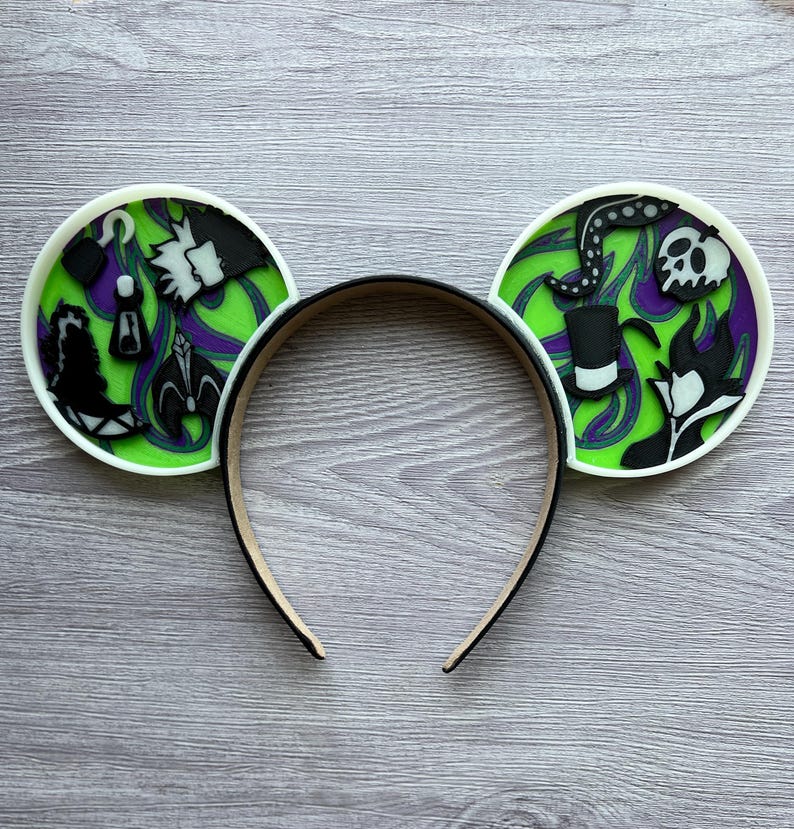 May include: A black headband with two round ear decorations. The ears feature a green and purple background with black and white character designs. The ears have a white border.