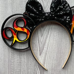 May include: Black headband with a black sequin bow and a black and red Mickey Mouse ear with the number 8 in a flame design.