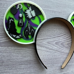 May include: Black headband with white trim and two round ear accents. The ear accents feature a green and purple design with black character silhouettes and a white border. The design includes a hook, a bottle, and a character's profile.