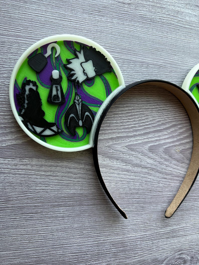 May include: Black headband with white trim and two round ear accents. The ear accents feature a green and purple design with black character silhouettes and a white border. The design includes a hook, a bottle, and a character's profile.