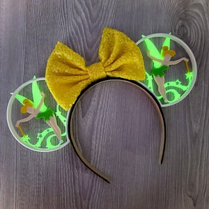 May include: A pair of white headband ears with a yellow bow. Each ear features a glow-in-the-dark Tinkerbell design with green wings and a wand.
