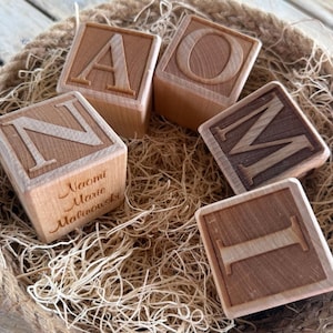 May include: Five wooden alphabet blocks with engraved letters A, O, M, I, and N. One block is engraved with the name "Naomi Marie Malinowski". The blocks are in a woven basket filled with straw.