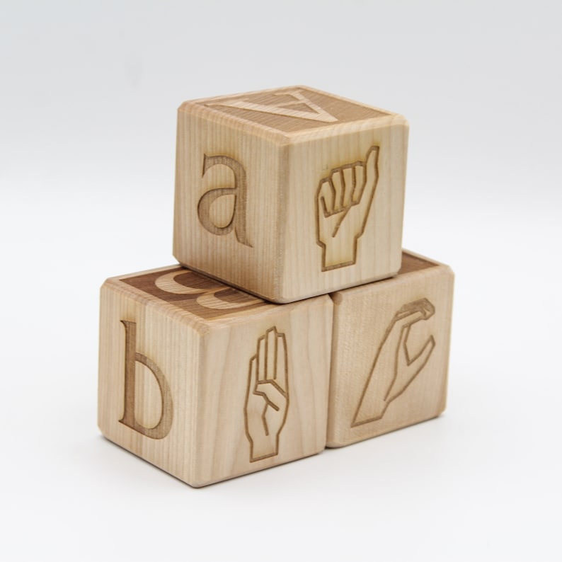 May include: Three wooden blocks with letters and sign language symbols. The top block has the letter "a" and a fist, the middle block has the letter "b" and a three-finger sign, and the bottom block has a "c" and a "c" sign.