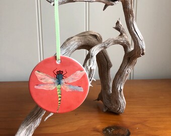 May include: A round, coral-colored ornament featuring a watercolor dragonfly design. The dragonfly has blue, green, and pink details. A light green ribbon is attached for hanging. The ornament is displayed against a natural wood background.
