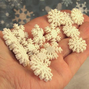 May include: A collection of small, off-white snowflake ornaments. The ornaments are held in a person's hand, showcasing their intricate, detailed designs. The snowflakes vary in size and shape, perfect for winter-themed crafts or decorations.