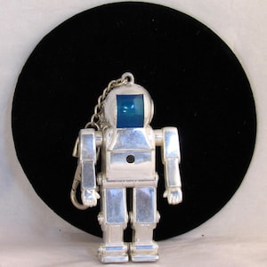 May include: A silver-colored robot keychain with a blue square face. The robot has articulated arms and legs, and a chain with a clasp. The robot is a vintage item.