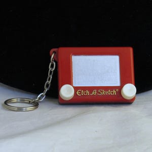 May include: A miniature red Etch A Sketch keychain featuring a rectangular frame, a white drawing screen, two white circular control knobs at the bottom, and the gold text Etch A Sketch printed on the front, attached to a silver metal ring.