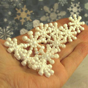 May include: Several white, pearl-like snowflake ornaments are displayed in a person's open hand. The snowflakes have a delicate, three-dimensional design, with multiple points radiating from a central hub. The background features a snowflake pattern.