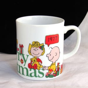 May include: White ceramic mug featuring a Christmas design with the text "Merry Christmas" in green. The mug has illustrations of cartoon characters, including a girl holding a teddy bear and a boy. A red speech bubble says "1977".