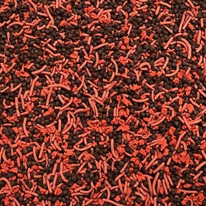 May include: Close-up of a textured surface covered in small, dark brown granules and scattered red, curved, and fragmented pieces. The contrasting colors and textures create a visually interesting pattern, possibly for food or decorative purposes.
