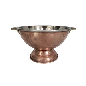 May include: A copper colander with a stainless steel interior and brass handles. The kitchen utensil has a pedestal base and is perforated with small holes for draining. The colander is designed for washing and straining food items.