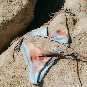 Puede incluir: Un conjunto de bikini de dos piezas con top y braguita anudados. La tela presenta un diseño tipo acuarela con tonos azul claro y marrón rojizo. El bikini se muestra sobre una superficie rocosa texturizada y de color claro.