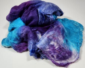 May include: A bundle of loose, unspun fiber featuring a vibrant gradient of colors ranging from deep royal purple to bright turquoise and light sky blue. The texture is soft and fibrous, with the colors blended in a marbled, hand-dyed pattern.