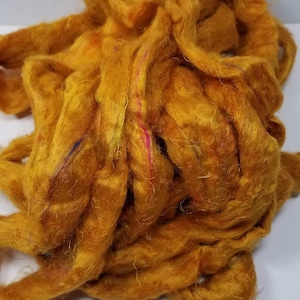 May include: A pile of golden yellow wool roving with subtle hints of orange and pink throughout. The roving is soft and fluffy, perfect for spinning or felting.