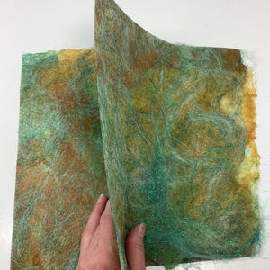 May include: A close-up of two sheets of handmade paper with a textured, fibrous surface. The paper features a blend of colors, including shades of green, brown, and yellow. The paper is being held open, revealing the layers and texture.