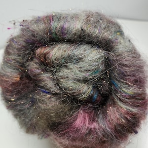 May include: A close-up of a ball of yarn with a mix of colors, including gray, purple, and teal. The yarn has a soft, fuzzy texture and is interspersed with strands of gold metallic thread, creating a shimmering effect. Ideal for knitting and crochet projects.