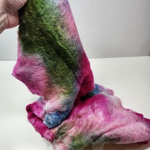 May include: A soft, fluffy, hand-dyed yarn in shades of pink, green, and blue. The yarn is folded and draped on a white surface.