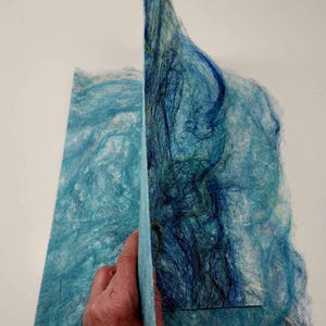 May include: Two sheets of handmade paper with a textured, layered appearance. The paper features shades of blue and teal, with darker blue fibers creating depth. The paper is held up, revealing the thickness and texture of the material.