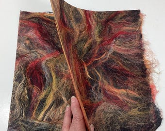 May include: A close-up of a felted textile with a mix of colors including red, brown, yellow, and black. The felted material is being lifted to reveal the layers and texture. The fibers are visible and create a unique, artistic appearance.