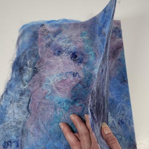 May include: A rectangular sheet of felted wool fiber featuring a blend of blue, purple, and white tones. The surface has a textured, fibrous appearance with swirling patterns of light blue and dark blue fibers layered over a purple base.