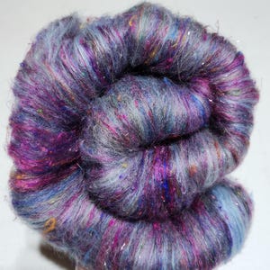 May include: A close-up of a skein of hand-dyed yarn. The yarn is a blend of purple, blue, and gray with flecks of other colors. It has a soft, textured appearance and a subtle shimmer. Ideal for knitting or crochet projects.