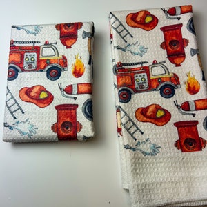 May include: Two white waffle-weave kitchen towels with a repeating pattern of red and orange fire trucks, fire hydrants, flames, and firefighter hats. One towel is folded, and the other is unfolded. The towels are on a white surface.
