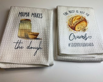 Op de afbeelding: Twee witte keukendoeken met wafelweefsel. Op de ene handdoek staat "Mama makes the dough" met een pot- en komafbeelding. De andere handdoek zegt "The best is yet to crumb" met een broodafbeelding en de hashtag #SOURDOUGHMAMA.