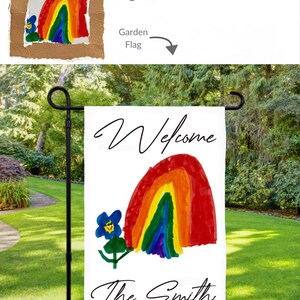 May include: A white garden flag featuring a rainbow and a blue flower design. The words "Welcome" and "The Smiths" are written in black script. The flag is displayed on a black metal stand in a garden setting.