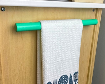 May include: A white waffle-weave kitchen towel with the text "Aiden's Kitchen" and utensil silhouettes. The towel hangs on a bright green wooden rod. The towel is part of a wooden play kitchen.