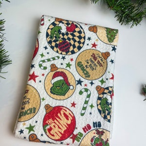 May include: Folded white Christmas towel with a festive Grinch design. The pattern includes Grinch-themed baubles, candy canes, and stars. The baubles feature the Grinch's face and text that reads "DWA TEST TURNS OUT I'm 100% GRINCH!!" and "THE GRINCH".