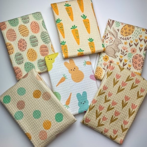 Pode incluir: Uma coleção de seis panos de cozinha dobrados com vários padrões temáticos de Páscoa. Os desenhos incluem ovos de Páscoa coloridos, cenouras, um coelho, Peeps, bolinhas e tulipas. Os panos são feitos de tecido texturizado.