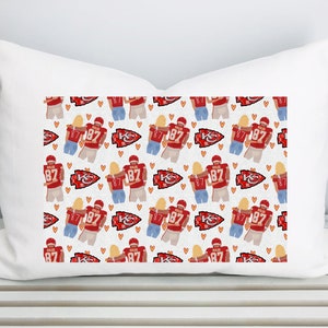 May include: White rectangular cushion featuring a repeating pattern of a football player in a red jersey with the number 87, a woman, hearts, and the team logo. A decorative item.