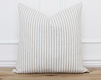 May include: A square pillow with a cream-colored background and vertical stripes in a muted beige color. The pillow is placed on a wooden surface against a white wall.