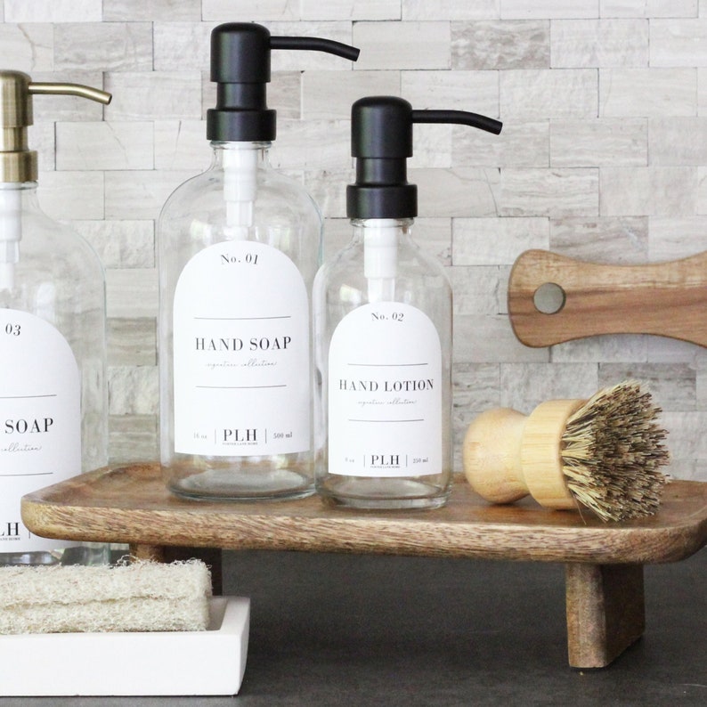 May include: Three glass bottles with black pump tops and white labels on a wooden shelf. The labels read "No. 01 Hand Soap", "No. 02 Hand Lotion", and "No. 03 Soap".