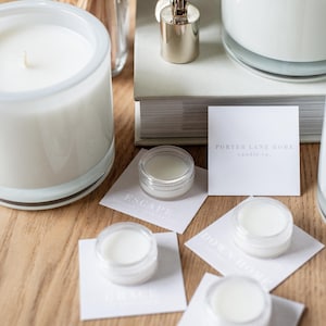 May include: A white candle in a clear glass jar with a white label that says "PORTER LANE HOME candle co.". There are several small clear jars with white lids on white cards with different scents written on them. The scents are "ESCAPE", "HOW TO", "ORANGE", and "CITRUS".