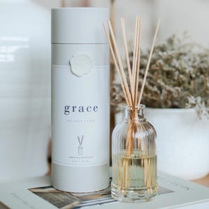 May include: A reed oil diffuser set with a cylindrical container and a glass bottle. The container is white with the word "grace" and "sea salt + sage" printed on it. The glass bottle contains a light yellow liquid and wooden reeds.