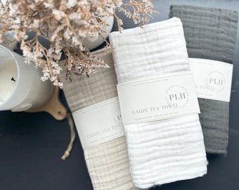 May include: Three folded gauze tea towels in neutral colors: beige, white, and gray. Each towel is wrapped with a white paper band that reads "GAUZE TEA TOWEL" and "PORTER LANE HOME". The towels are approximately 16 x 24 inches.